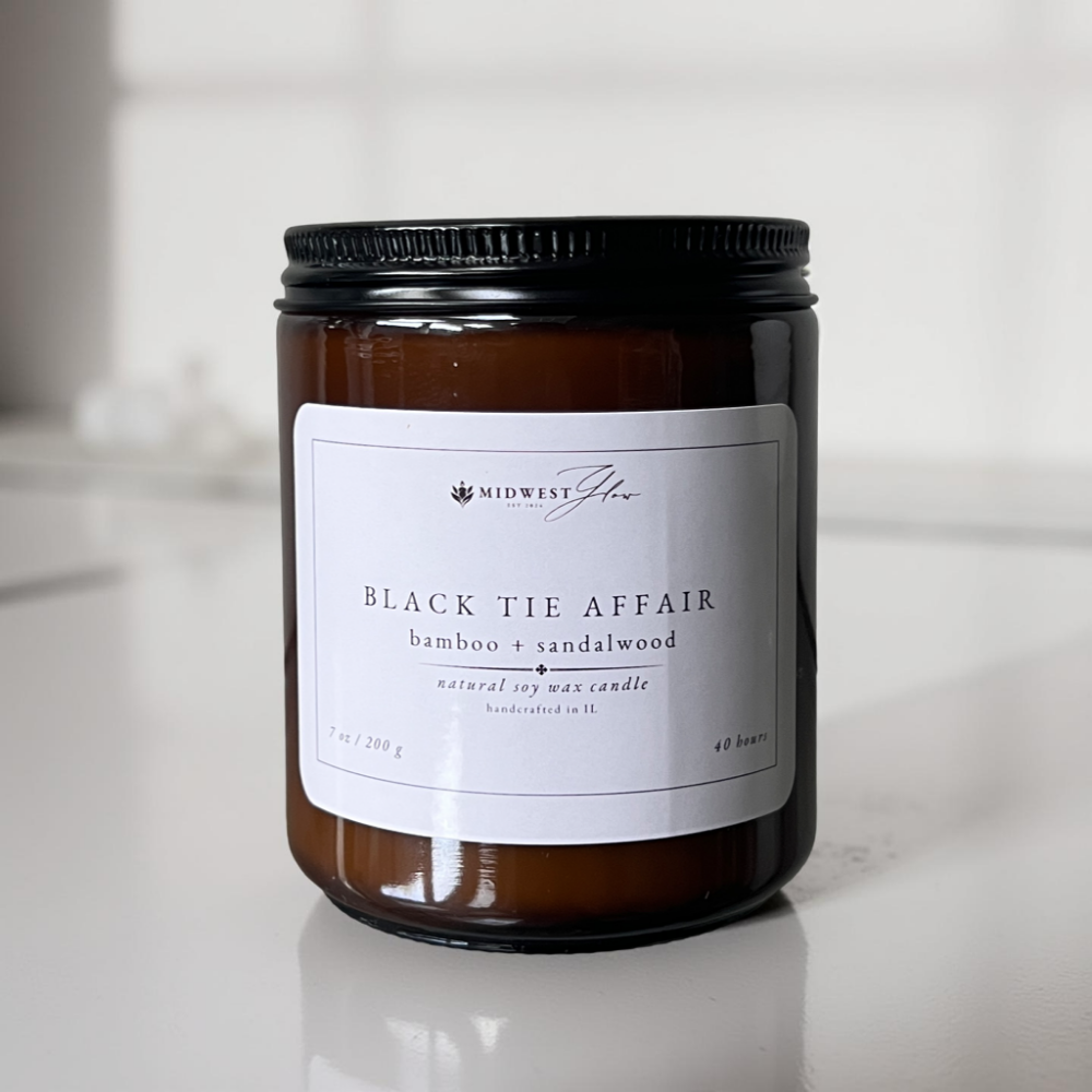 An amber glass candle jar sits in a pristine white photo setting with natural and studio lighting showcasing the Black Tie Affair scented soy candle by Midwest Glow