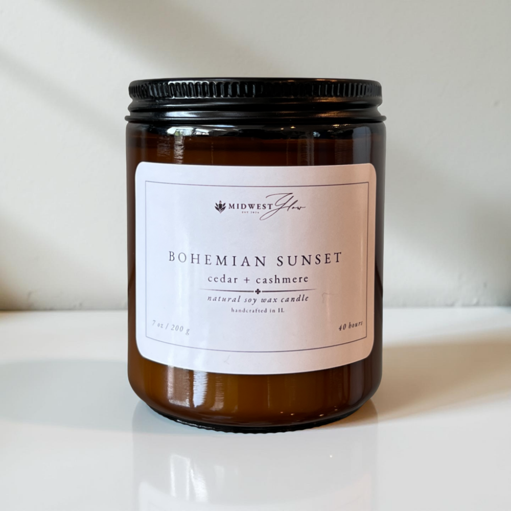 An amber glass candle jar sits in front of a pristine white setting with natural and studio lighting creating a beautiful ambience to showcase the Bohemian Sunset scented soy candle by Midwest Glow