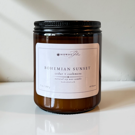 An amber glass candle jar sits in front of a pristine white setting with natural and studio lighting creating a beautiful ambience to showcase the Bohemian Sunset scented soy candle by Midwest Glow