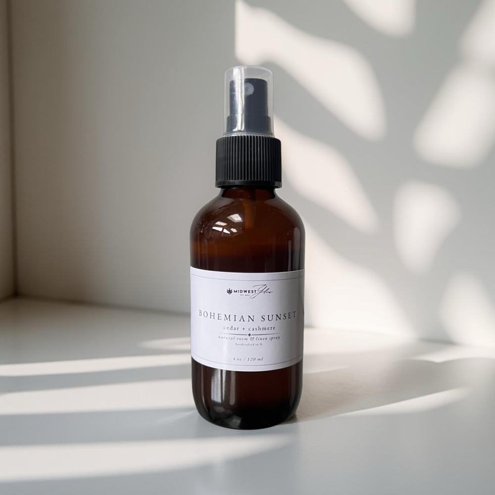 An amber glass spray bottle sits in a pristine white photo setting showcasing the Bohemian Sunset scented natural room and linen spray by Midwest Glow