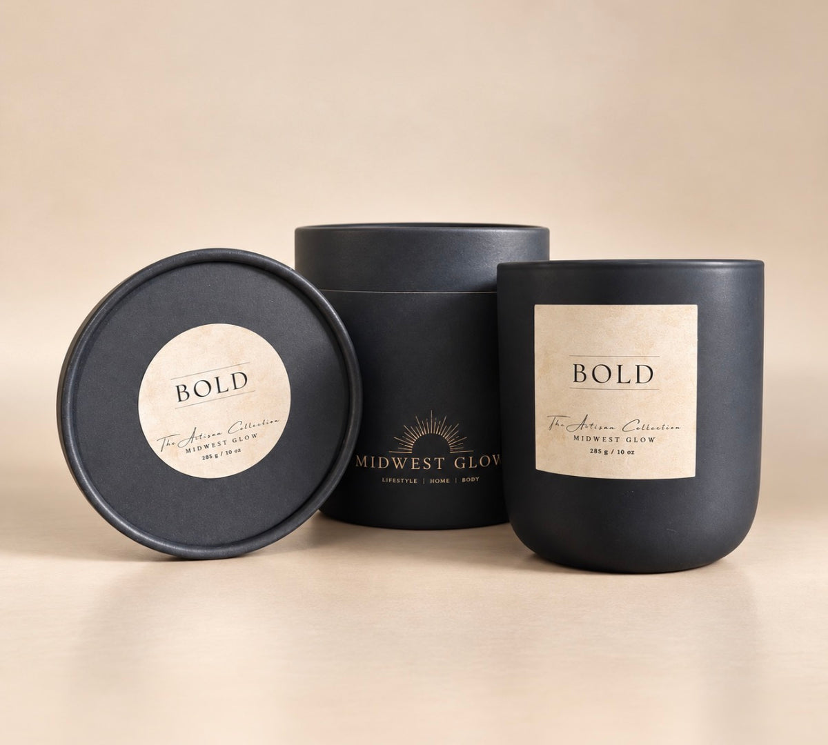 Three matte black candle packaging pieces on a warm beige studio background: a round lid with a cream label reading BOLD, a cylindrical tube with Midwest Glow branding, and a matching jar with a cream label.