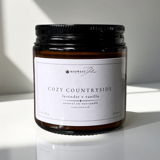 A 3 oz version of the Cozy Countryside candle by Midwest Glow sits in front of a pristine white wall backdrop with subtle shadows and lighting to enhance the products elegance