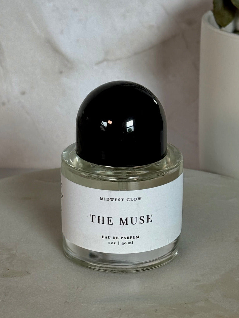 A clear glass perfume bottle with a black spherical cap and a white label reading 'THE MUSE' and 'MORWEIGHT GLOW', placed on a round stone surface with a neutral textured background and a potted plant in the upper right.
