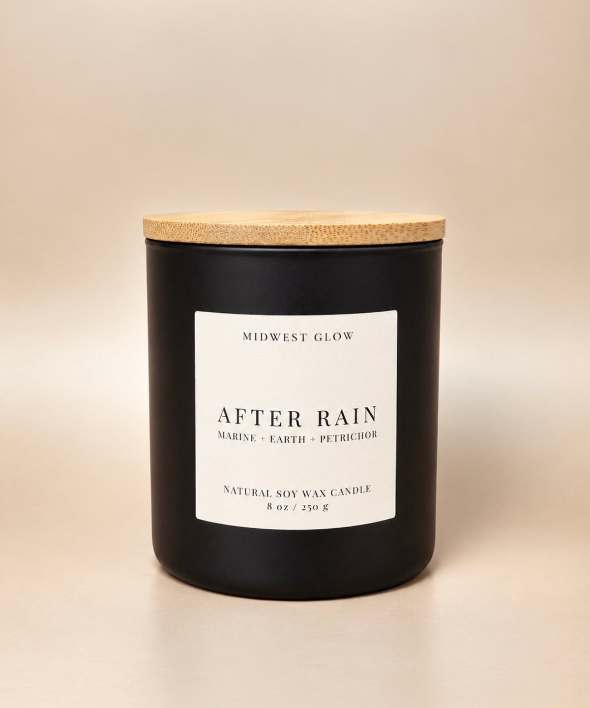 A matte black soy wax candle jar with a light wood lid and a white label reading 'AFTER RAIN' by Midwest Glow, photographed on a warm beige background.