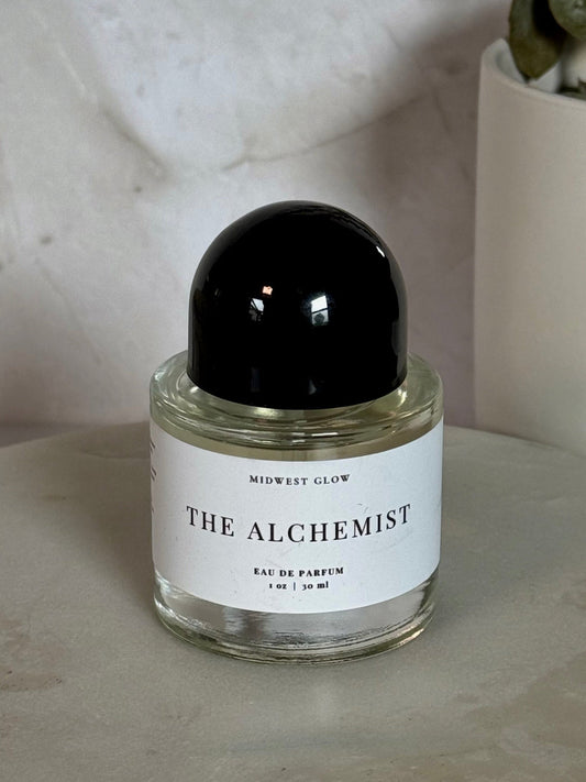 A small glass perfume bottle labeled 'THE ALCHEMIST' by Midwest Glow, with a glossy black cap and white label, set on a round stone surface with a neutral textured background and a potted plant in the corner.
