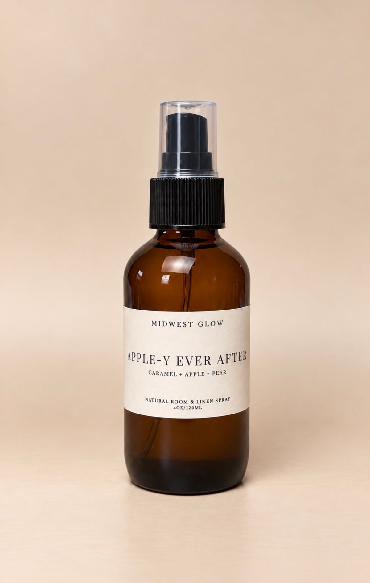 An amber glass spray bottle with a black pump and a white label reading 'Midwest Glow Apple-Y Ever After' on a warm beige background.