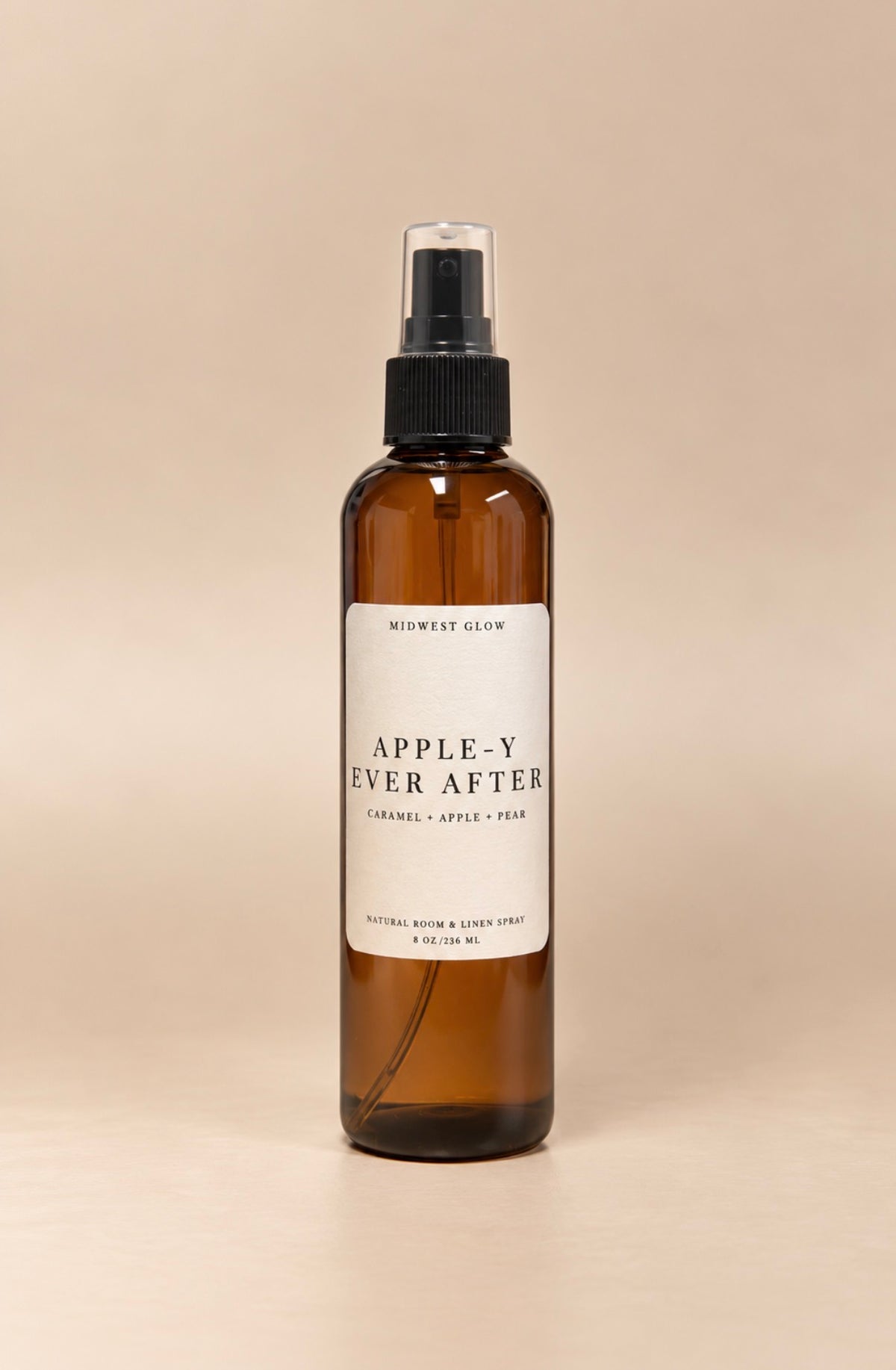 An amber glass room spray bottle with a black spray nozzle and a white label reading 'MIDWEST GLOW APPLE-Y EVER AFTER', set against a solid beige background.