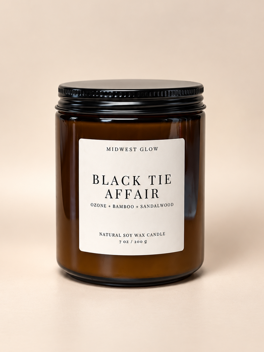 Midwest Glow Black Tie Affair natural soy wax candle in an amber glass jar with a black metal lid and a white label, set against a solid beige background.