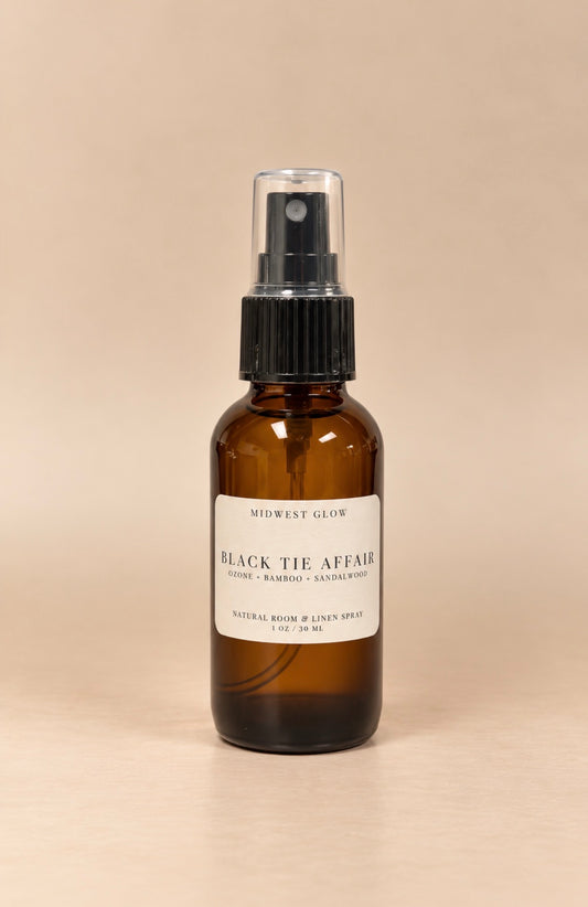 An amber glass room spray bottle with a black spray cap and a white label reading 'MIDWEST GLOW BLACK TIE AFFAIR'.