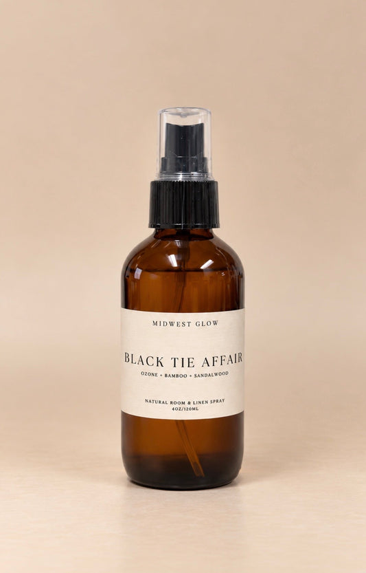 An amber glass spray bottle with a black pump and a white label reading 'MIDWEST GLOW BLACK TIE AFFAIR' on a warm beige background.