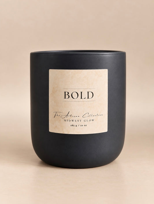 A matte black candle jar with a square cream label reading BOLD and The Artisan Collection, centered on a warm beige background.