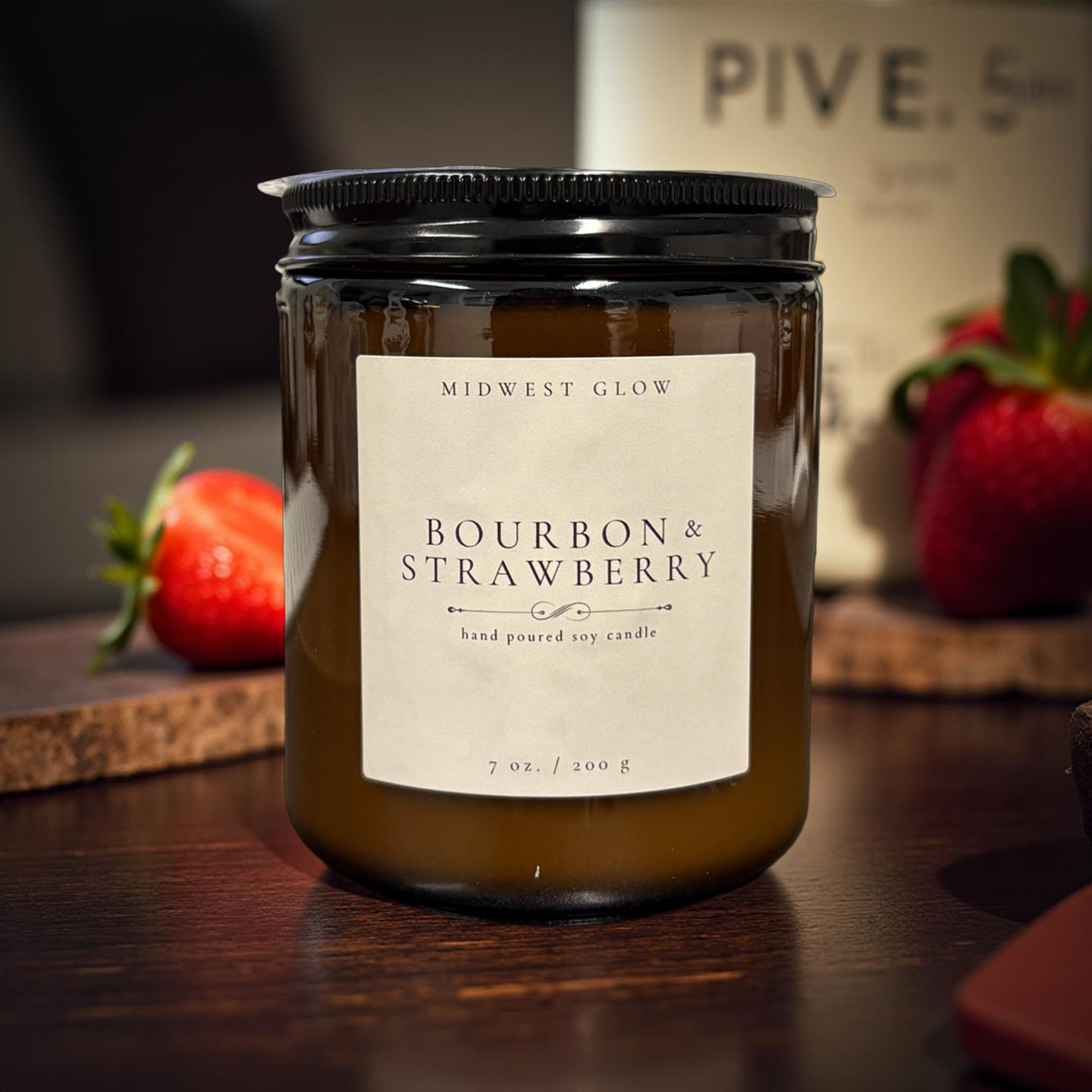 Candle labeled 'Bourbon & Strawberry' by Midwest Glow on a wooden surface with strawberries in the background.