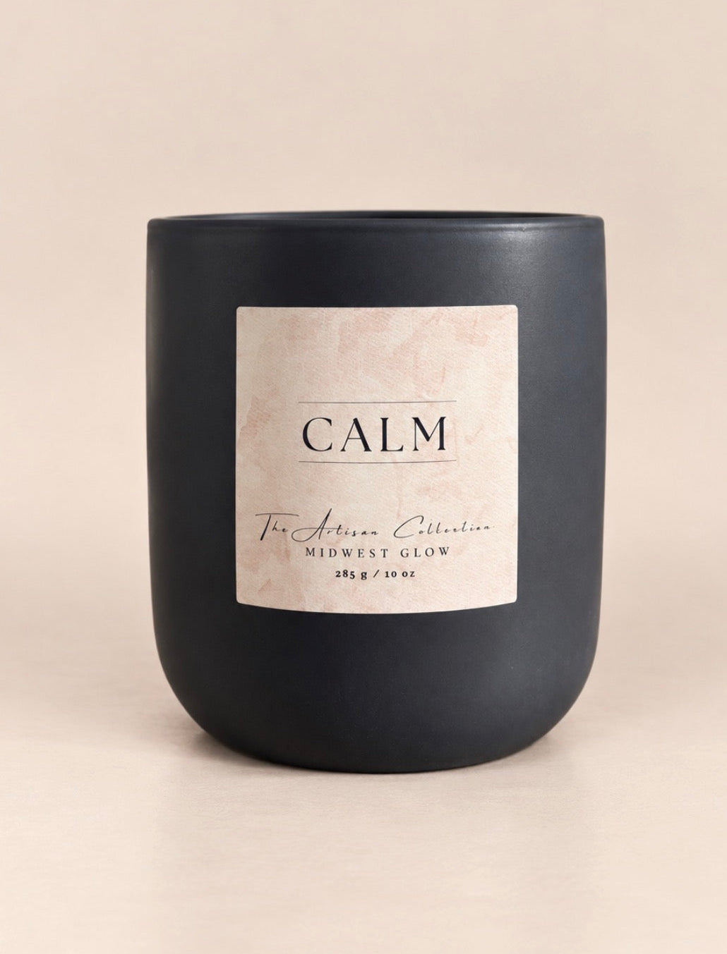 A matte black jar candle with a cream-colored label reading 'CALM' and 'Midwest Glow', set against a solid beige background.