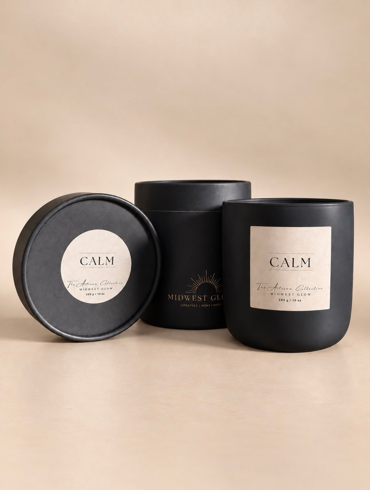 Three black candle packaging pieces on a warm beige background: a round lid with a silver label reading CALM, a cylindrical box with gold branding, and a matte black candle jar with a silver label reading CALM.