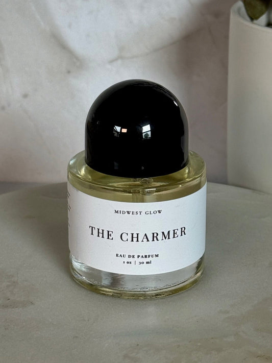 A small glass perfume bottle labeled 'THE CHARMER' by Mowest Glow, with a black spherical cap and pale yellow liquid, set on a light stone surface with a neutral textured background and a potted plant in the corner.