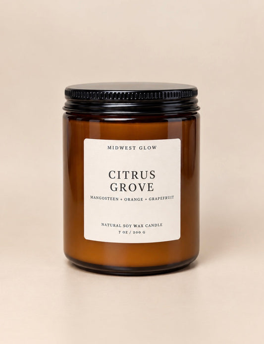 An amber glass jar candle with a black metal lid and a white label reading 'CITRUS GROVE' by Midwest Glow, centered on a warm beige background.