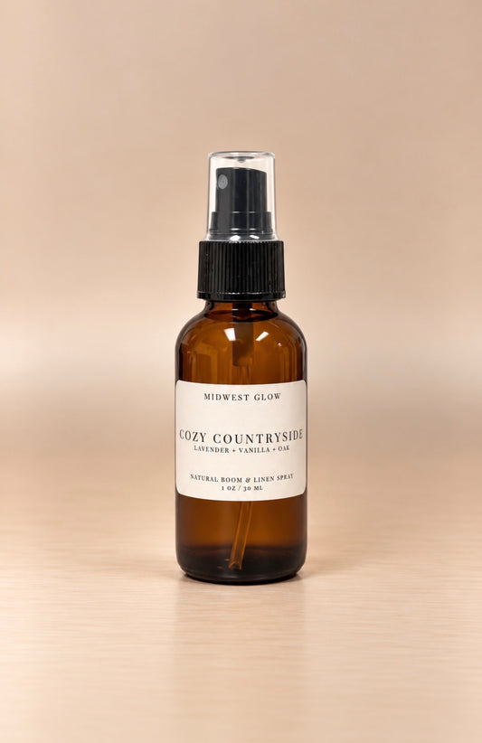 An amber glass spray bottle with a black pump and a white label reading 'Midwest Glow Cozy Countryside' on a light wood surface against a warm beige background.