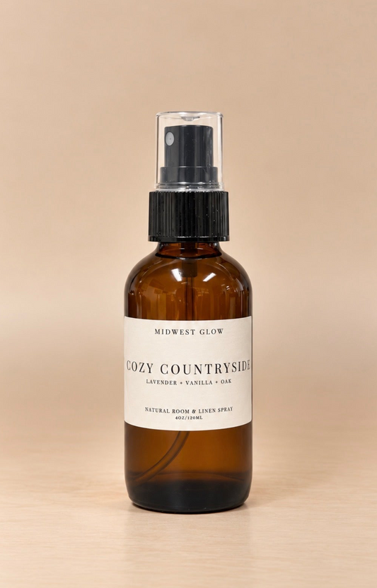 An amber glass room spray bottle with a black spray cap and a white label reading 'Midwest Glow Cozy Countryside', set against a warm beige background.