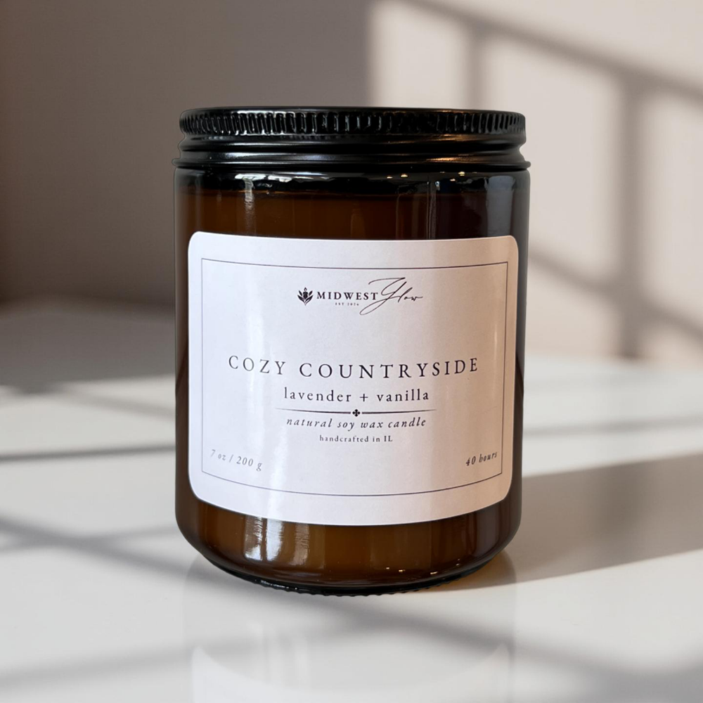 An amber glass candle jar sits in a pristine white photo studio with natural and studio lighting showcasing the Cozy Countryside scented soy candle by Midwest Glow