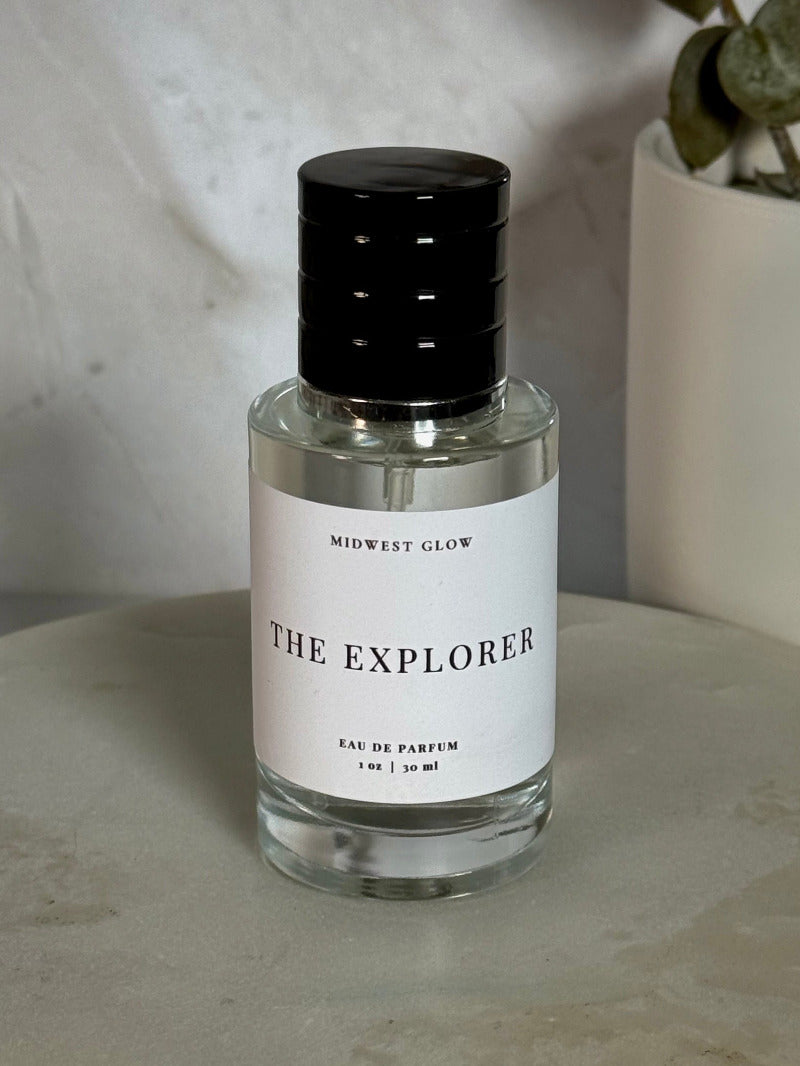 Midwest Glow 'The Explorer' Eau de Parfum in a clear glass bottle with a black cap and white label, set on a round stone tray with a neutral background and a potted plant.
