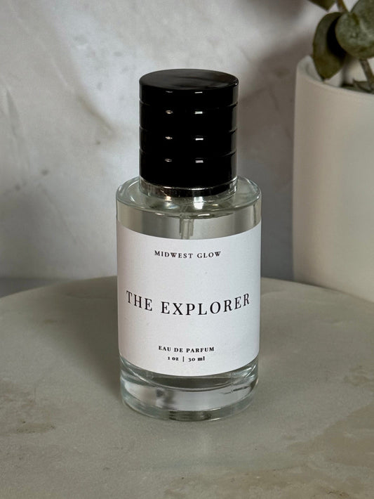 Midwest Glow 'The Explorer' Eau de Parfum in a clear glass bottle with a black cap and white label, set on a round stone tray with a neutral background and a potted plant.