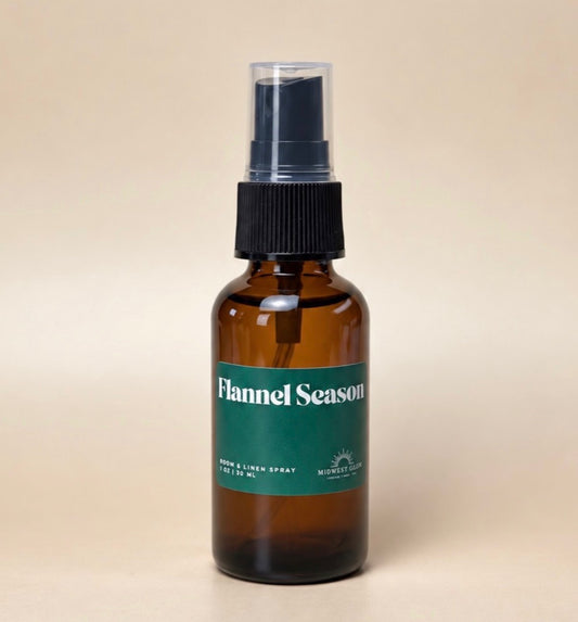 An amber glass spray bottle with a black pump and a green label reading 'Flannel Season' on a warm beige background.