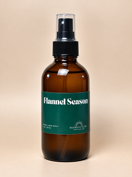 An amber glass spray bottle with a black pump and a dark green label reading 'Flannel Season' by Midwest Glow, centered on a warm beige background.