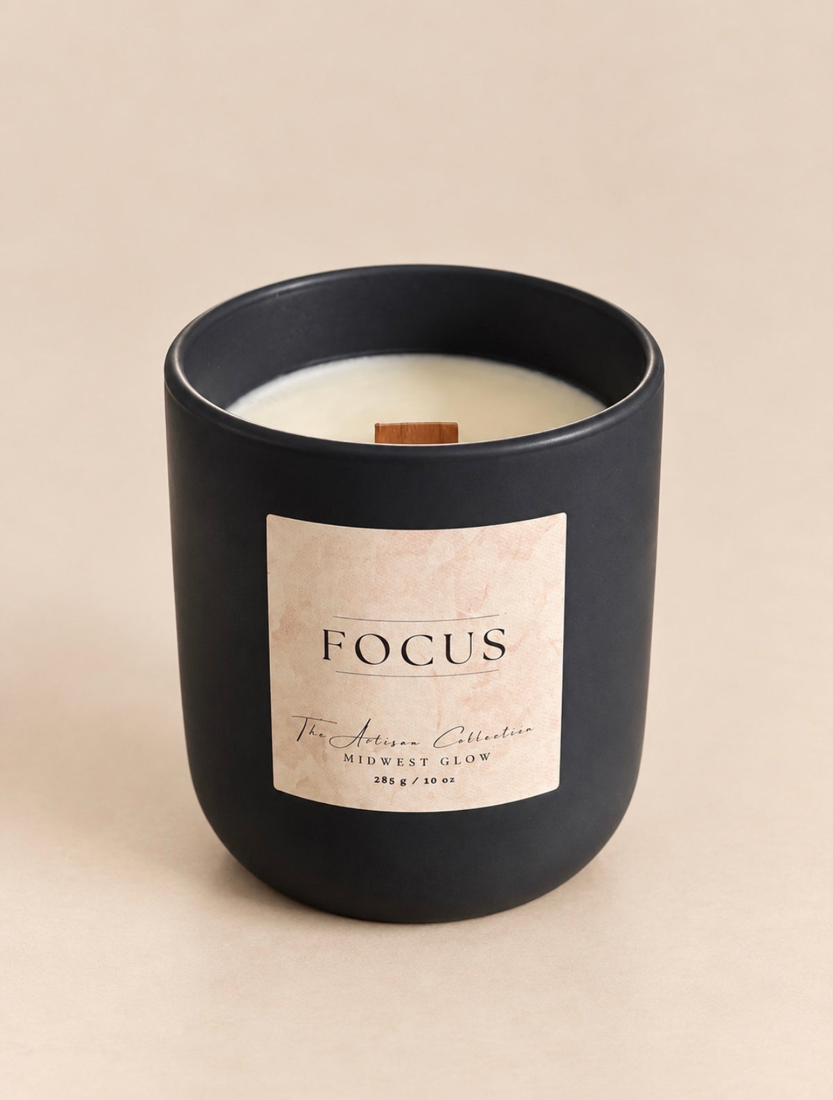 A matte black jar candle with a cream-colored wax and a single wooden wick, featuring a square beige label that reads 'FOCUS' and 'The Artisan Collection'.