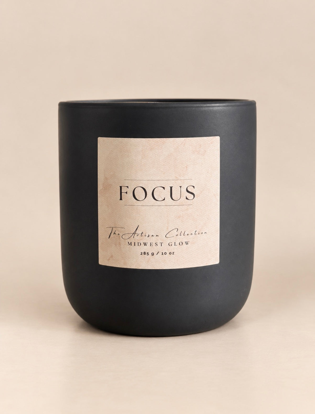 A matte black jar candle with a cream-colored rectangular label reading 'FOCUS' and 'The Artisan Collection', photographed on a warm beige background.