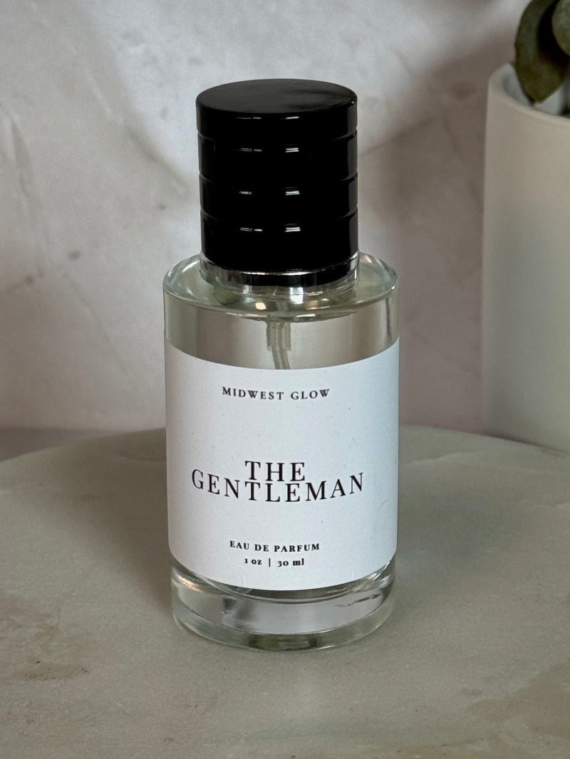 A clear glass bottle of MidWest Glow 'The Gentleman' Eau de Parfum with a black cap and white label, set on a round stone pedestal with a neutral background and a potted plant.