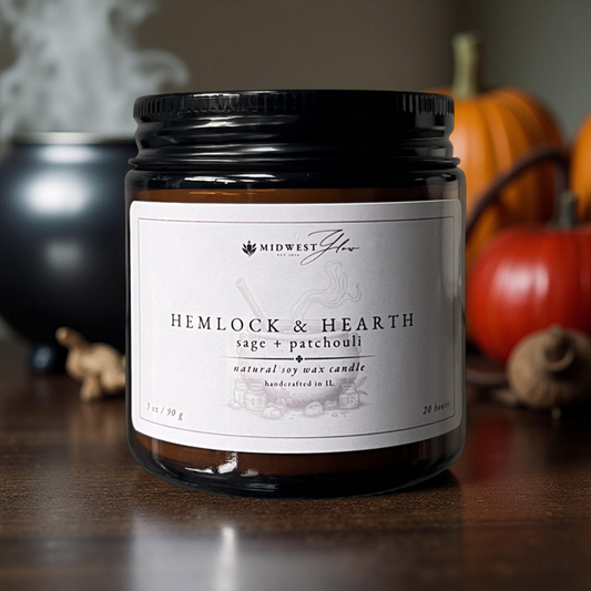 Candle jar labeled 'Hemlock & Hearth' with pumpkins and decorative elements in the background