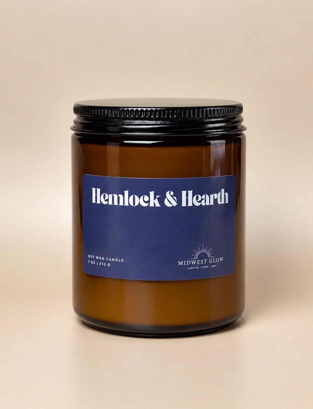 An amber glass soy wax candle jar with a black metal lid and a blue label reading 'Hemlock & Hearth' by Midwest Glow, set against a plain beige background.