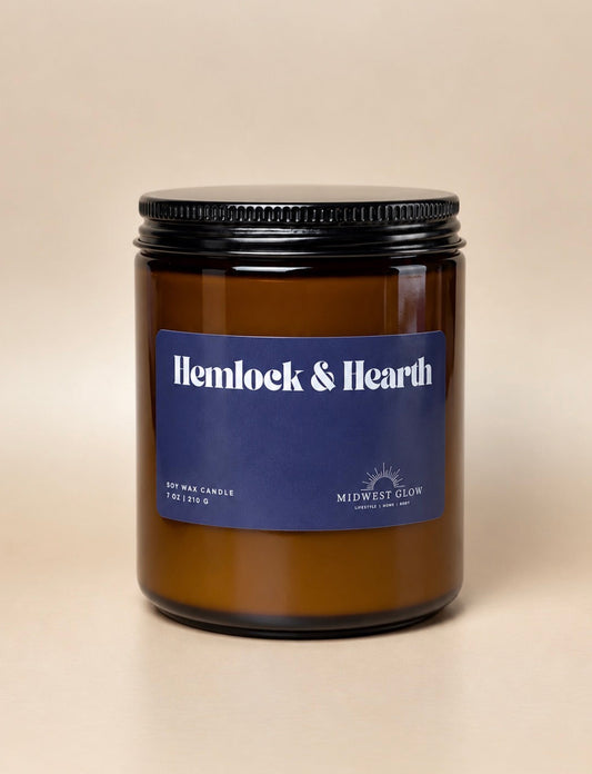 An amber glass soy wax candle jar with a black metal lid and a blue label reading 'Hemlock & Hearth' by Midwest Glow, set against a plain beige background.