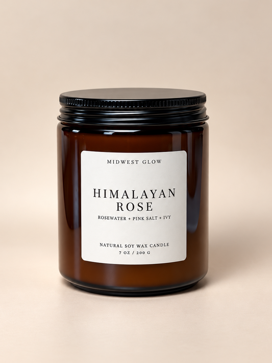 A Midwest Glow candle in an amber glass jar with a black lid and a white label reading 'HIMALAYAN ROSE' on a warm beige background.