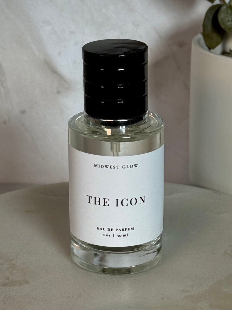 Midwest Glow 'The Icon' Eau de Parfum in a clear glass bottle with a white label and a black cap, set on a round marble tray with a neutral background and a potted plant.