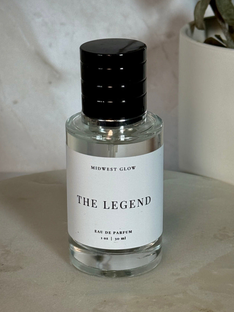 Midwest Glow 'The Legend' Eau de Parfum in a clear glass bottle with a black cap and white label, set on a stone surface next to a potted plant.