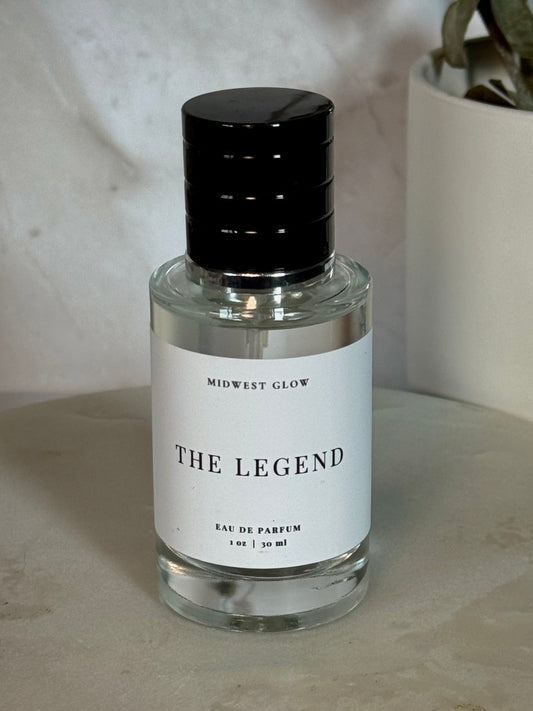 Midwest Glow 'The Legend' Eau de Parfum in a clear glass bottle with a black cap and white label, set on a stone surface next to a potted plant.