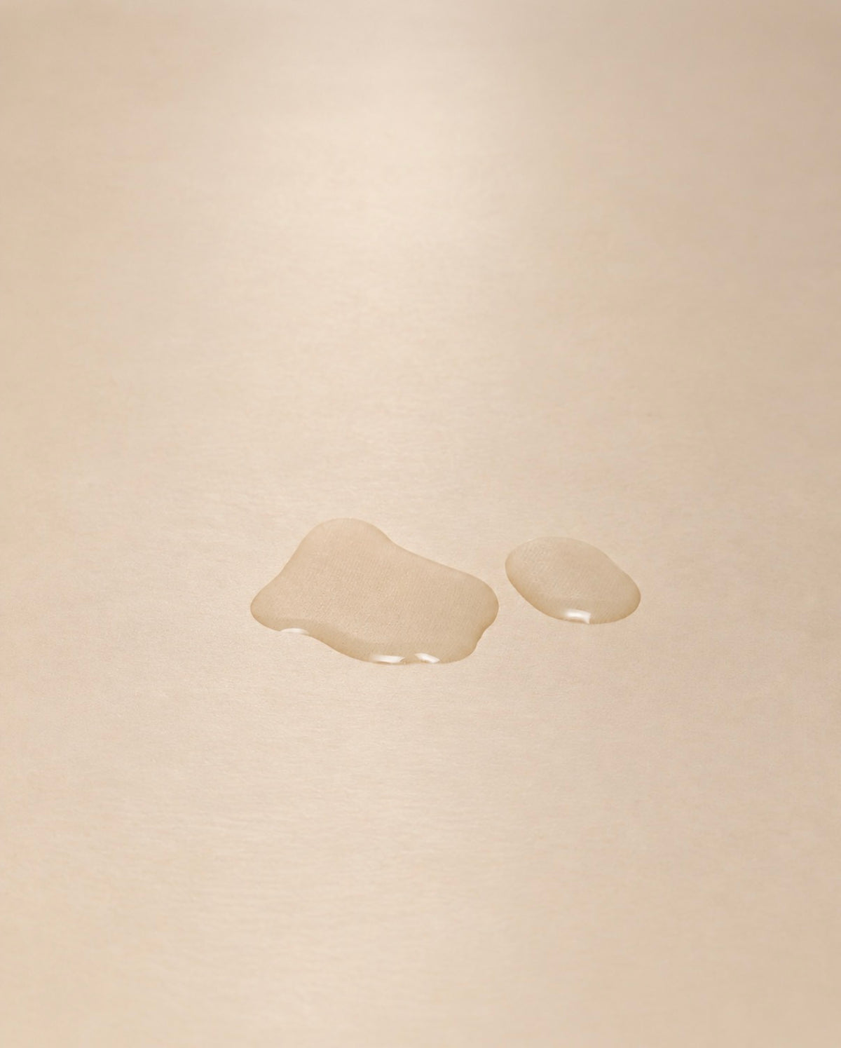 Two small translucent droplets of clear liquid resting on a smooth, warm beige surface.