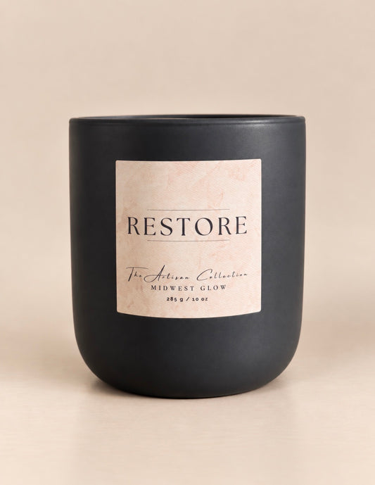 A matte black candle jar with a square cream label reading 'RESTORE' and smaller text 'For Artisan Collection' and 'MIDWEST GLOW', photographed on a warm beige background.
