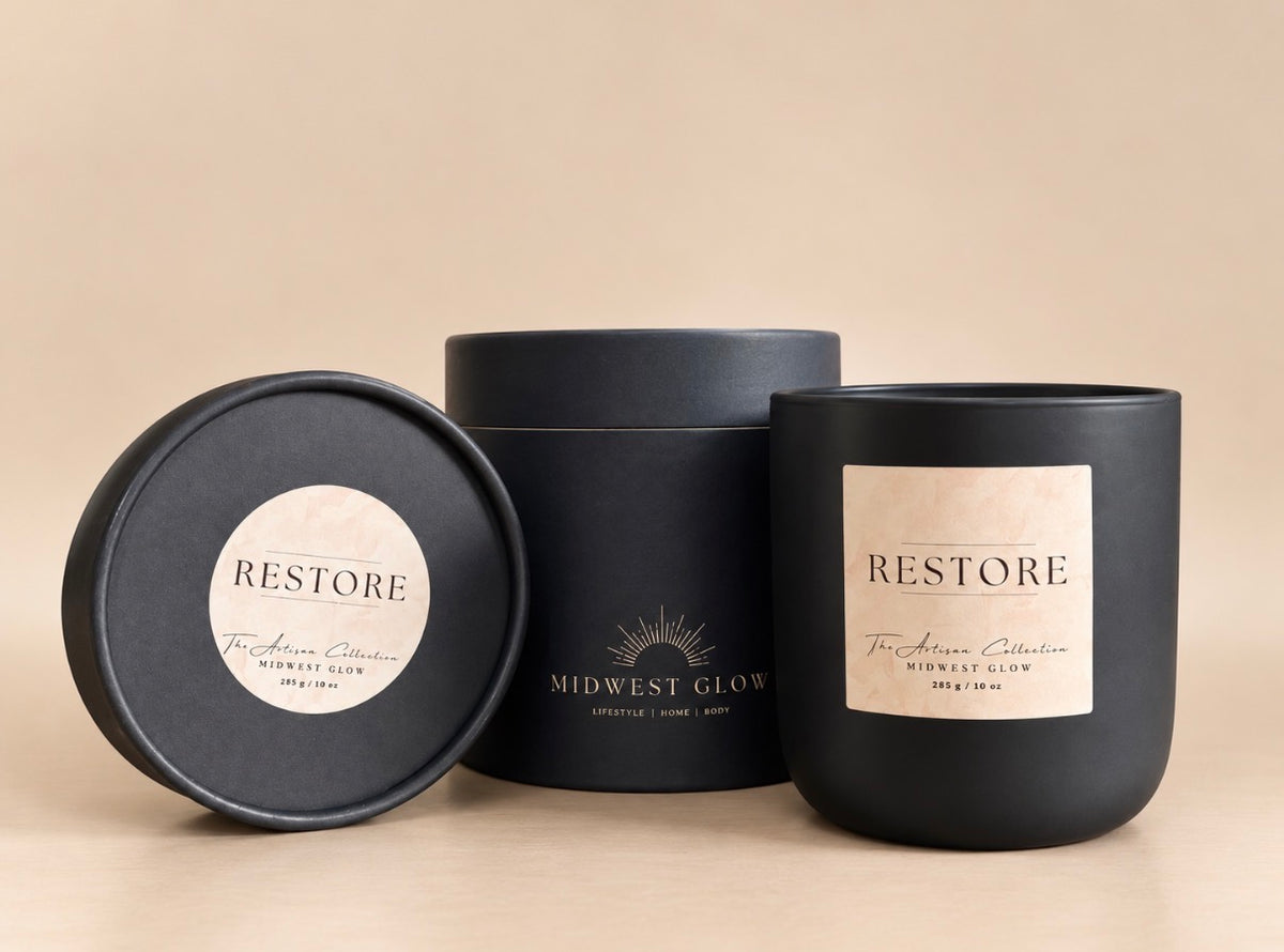 Three matte black candle packaging pieces on a warm beige background: a round lid with a circular Restore label, a cylindrical box with Midwest Glow branding, and a matching black candle vessel with a large Restore label.