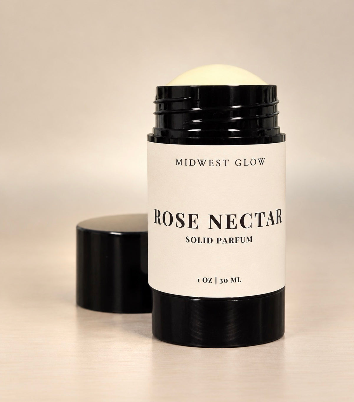 Midwest Glow Rose Nectar solid perfume stick in a black metal container with a white label, shown with the cap off and standing beside it on a light beige background.