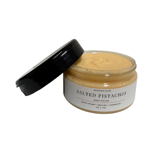 A jar of Midwest Glow Salted Pistachio Body Polish with its black lid leaning against the open container, set against a plain white background.