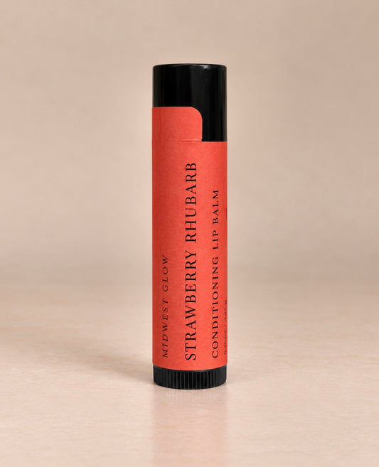 A single tube of lip balm standing upright on a light beige surface against a matching beige background. The tube has a black cap and base with a coral-red label that reads 'MIDWEST GLOW STRAWBERRY RHUBARB CONDITIONING LIP Balm'.