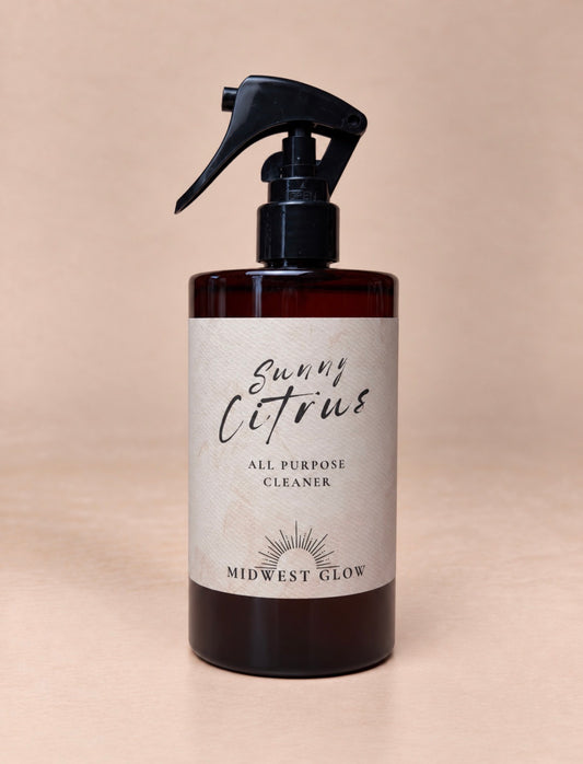 A dark amber glass spray bottle of Midwest Glow Sunny Citrus All Purpose Cleaner with a black trigger sprayer and a beige label, set against a solid beige background.