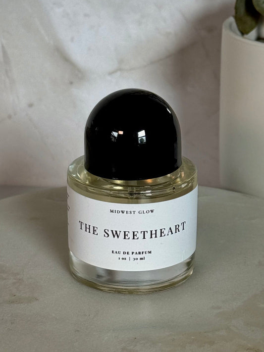 Midwest Glow 'The Sweetheart' Eau de Parfum in a clear glass bottle with a black cap and white label, styled on a marble tray with a potted plant in the background.