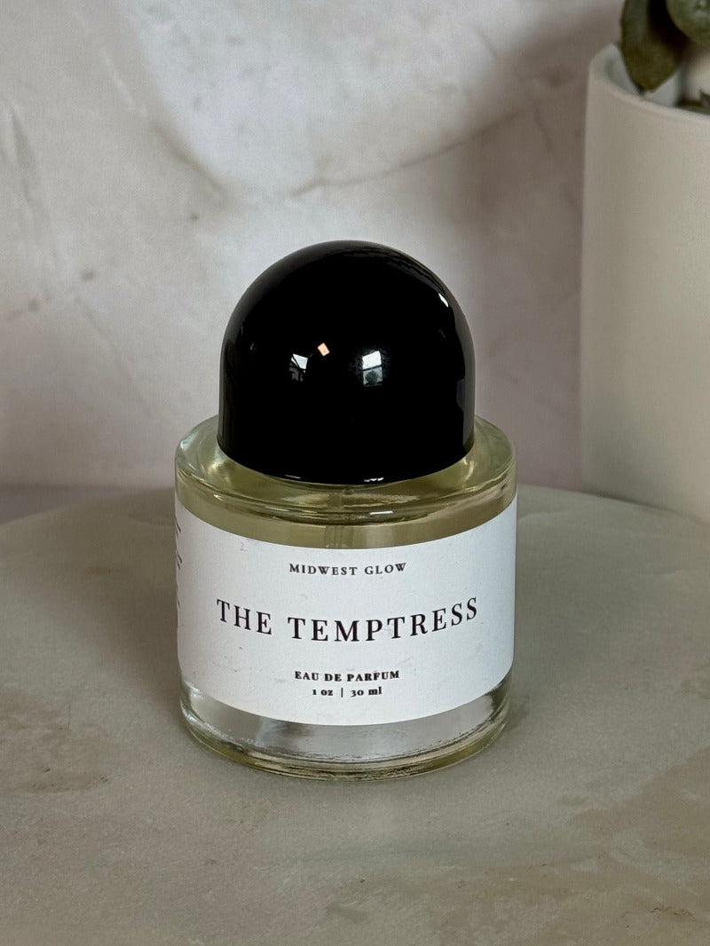 Midwest Glow 'The Temptress' Eau de Parfum in a clear glass bottle with a black spherical cap and a white label, displayed on a round stone slab with a neutral background.