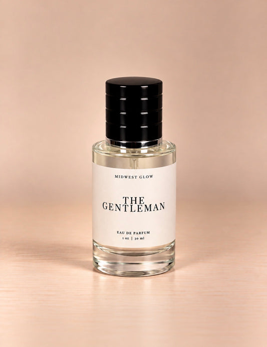 A clear glass bottle of Eau de Parfum with a white label reading 'THE GENTLEMAN' and 'MIDWEST GLOW', topped with a black ribbed cap, set on a light wood surface against a warm beige background.