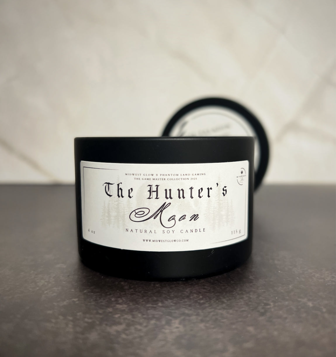 A candle in a black tin showing the label that reads The Hunter's Moon by the company Midwest Glow