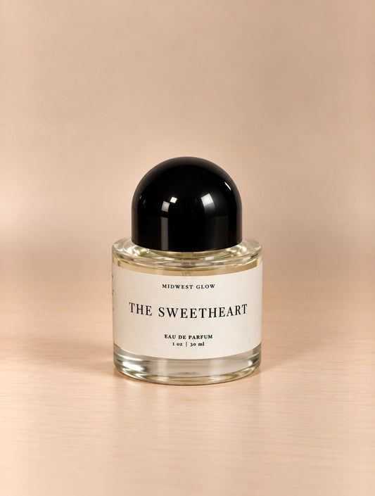 A 1 oz glass bottle of Midwest Glow 'The Sweetheart' Eau de Parfum, featuring a black dome cap and a cream-colored label, set against a warm beige background.