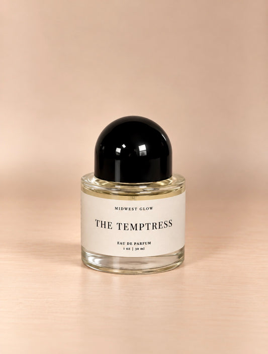 Midwest Glow The Temptress Eau De Parfum in a clear glass bottle with a black spherical cap and a white label, set against a warm beige background.